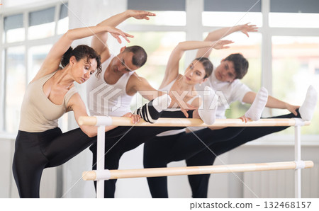 Group of young female and male ballet dancers learn grand batman position near ballet barre in gym Group of young female and male ballet dancers learn grand batman position near ballet barre in gym 132468157