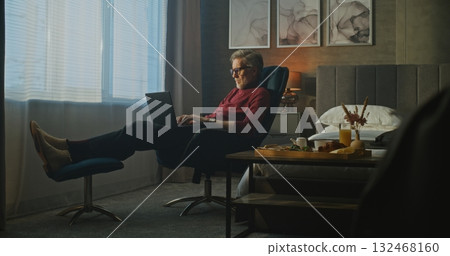 Gray Haired Man Sits Relaxed in Hotel Chair, Typing on Laptop in Morning Light Gray Haired Man Sits Relaxed in Hotel Chair, Typing on Laptop in Morning Light 132468160