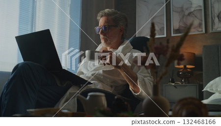 Man in Hotel Bathrobe Sits by Window With Laptop and Breakfast, Focused on Work. Man in Hotel Bathrobe Sits by Window With Laptop and Breakfast, Focused on Work. 132468166