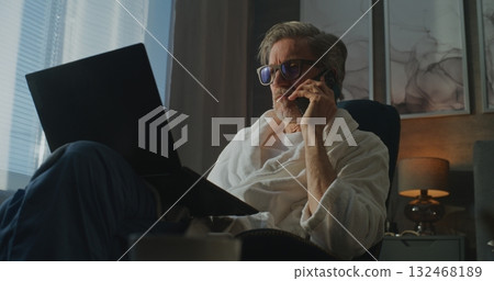 Professional Man in Bathrobe With Glasses Talks on Smartphone and Use Laptop 132468189