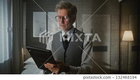 Gray Haired Man in Vest and Tie Stands, Looking Down at Digital Tablet 132468194