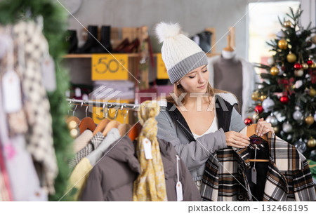 Girl client looks at products in store, looks closely at checkered coat, picks up wardrobe item Girl client looks at products in store, looks closely at checkered coat, picks up wardrobe item 132468195