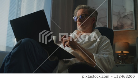 Stylish Gray Haired Man in White Bathrobe Checks Emails on Laptop, Enjoying Morning Light and Coffee 132468200