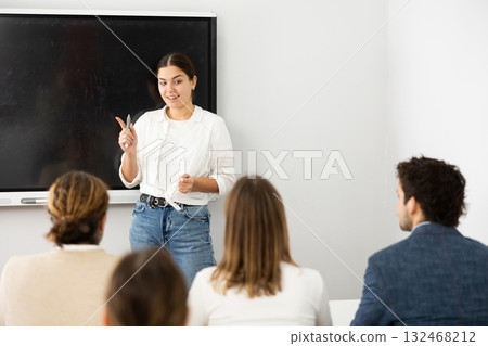 Young female teacher near interactive board, gesturing with hand in front of people sitting at desks in auditory Young female teacher near interactive board, gesturing with hand in front of people sitting at desks in auditory 132468212