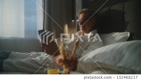 Man Enjoys Enjoys Breakfast in Bed and Phone in Hotel Room 132468217