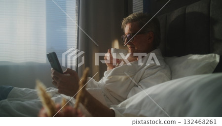 Man Enjoys Enjoys Breakfast in Bed and Phone in Hotel Room 132468261
