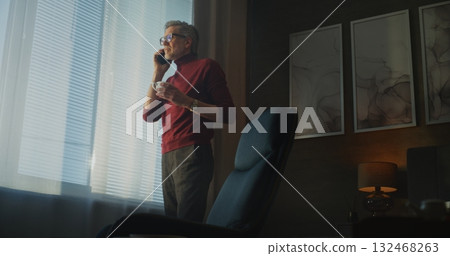 Confident Businessman With Gray Hair and Glasses Drinks Coffee Near Window While Making Phone Call 132468263