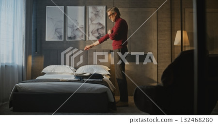 Gray Haired Mature Man in Red Turtleneck Packs His Belongings Into Suitcase in Stylish Hotel Room Gray Haired Mature Man in Red Turtleneck Packs His Belongings Into Suitcase in Stylish Hotel Room 132468280