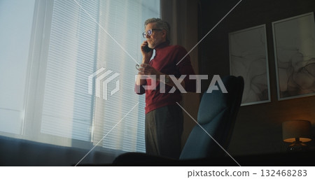 Mature Man in Red Turtleneck and Glasses Stands by Large Window, Drinking Coffee 132468283
