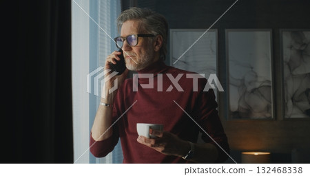 Stylish Mature Man in Red Turtleneck and Glasses Stands By Panoramic Window in Bright Hotel Room Stylish Mature Man in Red Turtleneck and Glasses Stands By Panoramic Window in Bright Hotel Room 132468338