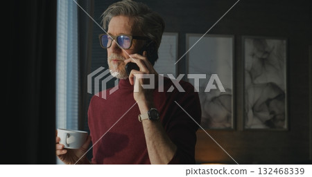 Elegant Gray Haired Man in Modern Hotel Room Holds Coffee Cup and Talks on Smartphone Elegant Gray Haired Man in Modern Hotel Room Holds Coffee Cup and Talks on Smartphone 132468339