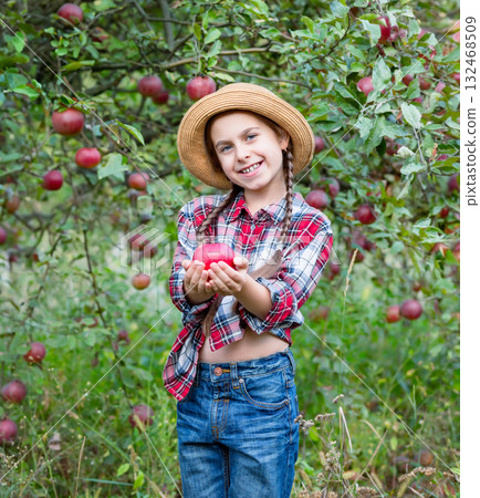 Cheerful girl stands on background an apple orchard with red apples, tastes, enjoys snacks, vitamins, 132468509