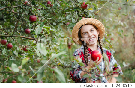 Cheerful girl stands on background an apple orchard with red apples, tastes, enjoys snacks, vitamins, organic fruits. She is dressed in checkered shirt and hat. Cheerful girl stands on background an apple orchard with red apples, tastes, enjoys snacks, vitamins, organic fruits. She is dressed in checkered shirt and hat. 132468510