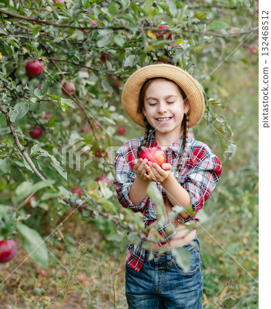 Cheerful girl stands on background an apple orchard with red apples, tastes, enjoys snacks, 132468512