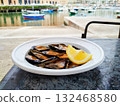 Plate of mussels with a lemon wedge served on a table by the waterfront in Bari, Italy. Background features docked boats on calm water under a partly cloudy sky, and buildings 132468580