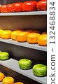 Assortment of cheeses displayed on gray shelves. Top shelf holds red wax-covered rounds, middle has orange rounds, and bottom features green wax-covered rounds. Netherlands, Holland cheese  132468834