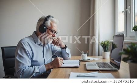 Senior Man Working from Home with Headphones, Laptop, and Notebook on Wooden Table Senior Man Working from Home with Headphones, Laptop, and Notebook on Wooden Table 132470110