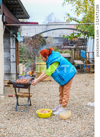 A 50-year-old Russian woman in a rural village grills marinated chicken outdoors, tending skewers over an open flame on a cool autumn day in Russia. A 50-year-old Russian woman in a rural village grills marinated chicken outdoors, tending skewers over an open flame on a cool autumn day in Russia. 132475286