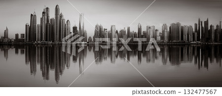 Monochrome city skyline with tall skyscrapers reflecting on calm water under a cloudy sky, Dubai Marina bay UAE 132477567