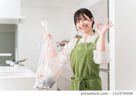A woman in an apron collecting trash in the trash can in the kitchen, a housewife A woman in an apron collecting trash in the trash can in the kitchen, a housewife 132478099
