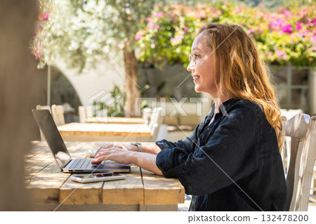 Side View of Smiling Woman Working on Laptop Outdoors in Cafe with Greenery and Natural Light 132478200