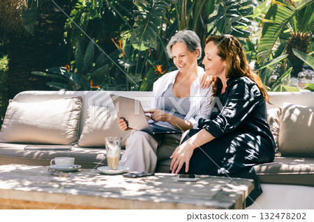 Two mature women happily working on a laptop while sitting on a sofa in a lush tropical garden. Two mature women happily working on a laptop while sitting on a sofa in a lush tropical garden. 132478202