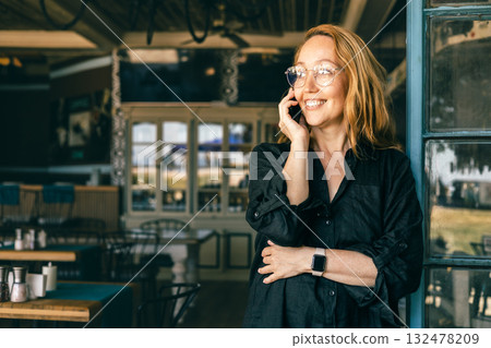 Happy Woman Talking on Phone Outside a Cozy Cafe, Wearing Glasses and Smartwatch 132478209