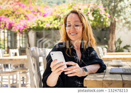 Smiling Woman Using Smartphone at Outdoor Cafe Table with Laptop and Coffee on a Sunny Day Smiling Woman Using Smartphone at Outdoor Cafe Table with Laptop and Coffee on a Sunny Day 132478213