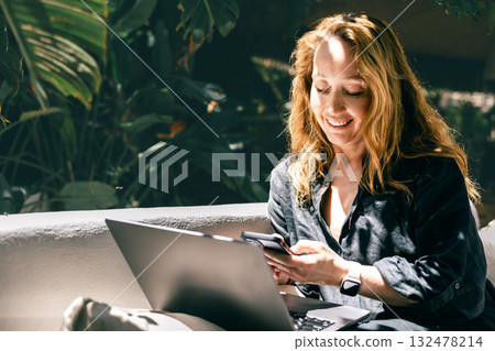 Cheerful woman using smartphone and laptop while working outdoors in a tropical environment 132478214