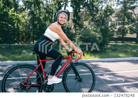 Happy active senior woman in helmet riding red bicycle on sunny path in green urban park 132478248