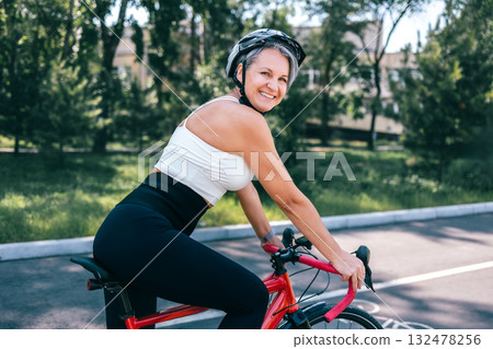 Confident mature woman smiling on red bicycle in activewear and helmet on sunny park bike lane 132478256