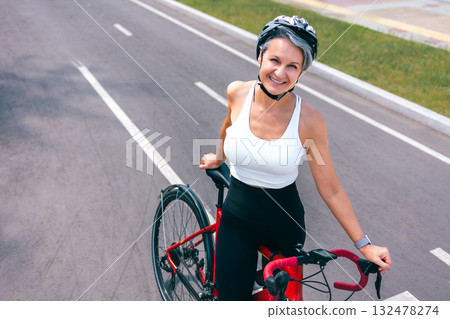 Happy Senior Woman in Helmet Standing with Bicycle on City Road in Sportswear and Smiling 132478274