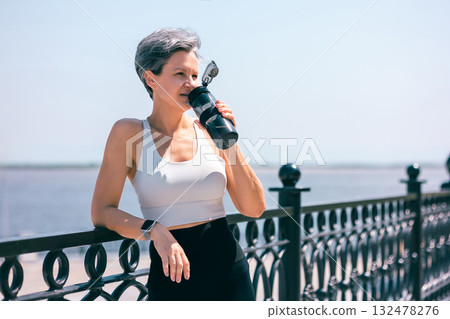 Active Woman Hydrating with Water Bottle by Waterfront After Outdoor Exercise Session 132478276