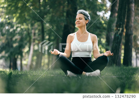 Smiling Mature Woman Meditating in Sukhasana Pose in Peaceful Forest for Mindful Wellness 132478290