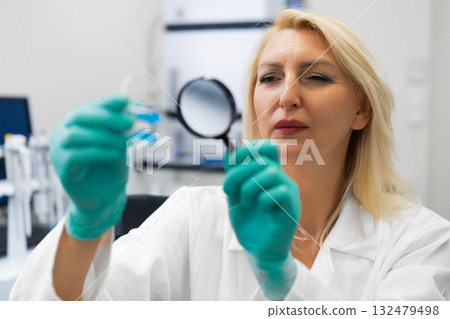 Blonde female chemist in lab using magnifying glass to study solutions in test tubes. 132479498