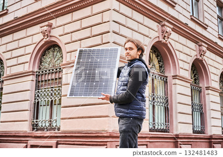 Man holding photovoltaic solar panel in front of historical building. Concept of integration of sustainable renewable energy sources into architecture. Man holding photovoltaic solar panel in front of historical building. Concept of integration of sustainable renewable energy sources into architecture. 132481483