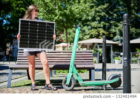 Happy woman charging her electric scooter from solar panel. Young female holding solar battery and catching sunlight. Concept of renewable energy resources. Happy woman charging her electric scooter from solar panel. Young female holding solar battery and catching sunlight. Concept of renewable energy resources. 132481494