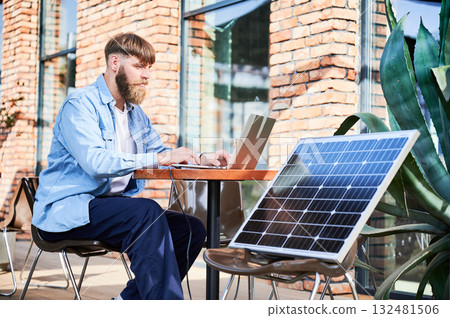 Man works on laptop at outdoor round table, with glass of iced coffee. Solar panel nearby, emphasizing sustainable, modern eco-friendly workspace that combines technology and sustainability. Man works on laptop at outdoor round table, with glass of iced coffee. Solar panel nearby, emphasizing sustainable, modern eco-friendly workspace that combines technology and sustainability. 132481506