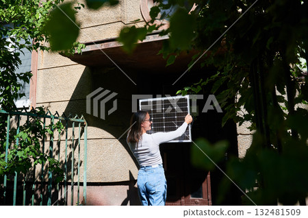 Woman holding photovoltaic solar panel in front of historical building, wearing plaid shirt and sunglasses. Concept of integration of sustainable renewable energy sources into old architecture. 132481509