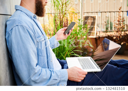 Bearded man works on laptop on lap and smartphone. Solar panel nearby, emphasizing sustainable, modern eco-friendly workspace that combines technology and sustainability. Bearded man works on laptop on lap and smartphone. Solar panel nearby, emphasizing sustainable, modern eco-friendly workspace that combines technology and sustainability. 132481510