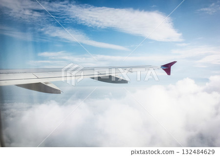 The image shows an airplane wing viewed from a passenger window seat. The sky is partially cloudy with blue sky visible, and sunlight is illuminating the scene, suggesting it could be daytime. The The image shows an airplane wing viewed from a passenger window seat. The sky is partially cloudy with blue sky visible, and sunlight is illuminating the scene, suggesting it could be daytime. The 132484629