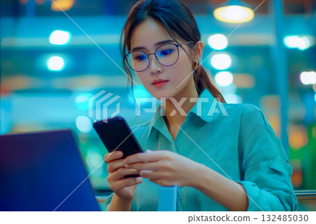 A woman with glasses focuses on her smartphone as she sits at a laptop in a lit cafe during the evening 132485030