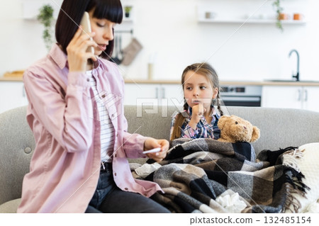 A concerned mother talks on the phone while checking her daughter's temperature, who is wrapped in a blanket on the couch, looking unwell. 132485154