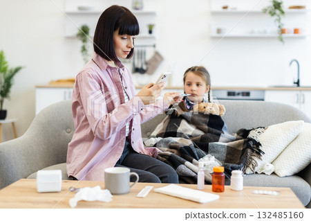 A concerned mother checks her daughter's temperature while she is wrapped in a blanket on the sofa, with medicine on the table. A concerned mother checks her daughter's temperature while she is wrapped in a blanket on the sofa, with medicine on the table. 132485160