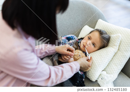 A concerned mother checks her sick daughter's temperature with a thermometer while she rests on a sofa, showing care and support. 132485180