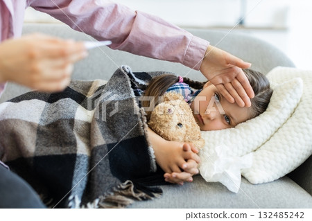 A concerned parent checks the temperature of a sick young girl with a thermometer while she rests on a couch, wrapped in a blanket. A concerned parent checks the temperature of a sick young girl with a thermometer while she rests on a couch, wrapped in a blanket. 132485242