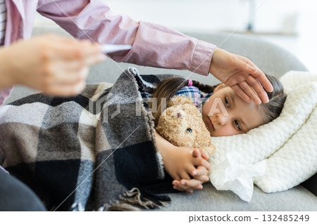 A young girl lies on a couch, wrapped in a blanket, while her mother checks her forehead for a fever. A teddy bear is held close. A young girl lies on a couch, wrapped in a blanket, while her mother checks her forehead for a fever. A teddy bear is held close. 132485249