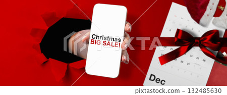 Human hand through hole paper showing Christmas big sale text on cellphone screen with red ribbon. Copy space. Christmas sale concept Human hand through hole paper showing Christmas big sale text on cellphone screen with red ribbon. Copy space. Christmas sale concept 132485630