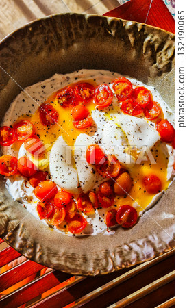 A plate of scrambled eggs topped with sliced cherry tomatoes. The dish is garnished with black pepper and served on a textured ceramic plate. 132490065