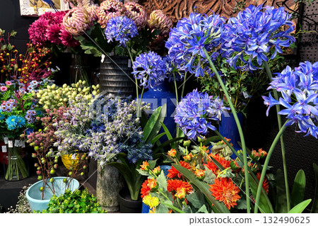 Large retail display of artificial flowers including blue agapanthus, purple thistle, and orange dahlias. Maximalist decor, color-saturated styling, faux floral diversity, seasonal botanical textures Large retail display of artificial flowers including blue agapanthus, purple thistle, and orange dahlias. Maximalist decor, color-saturated styling, faux floral diversity, seasonal botanical textures 132490625